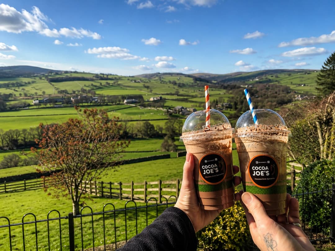 Two iced Cocoa Joe's drinks held against a wide blue-sky dale view in Nidderdale