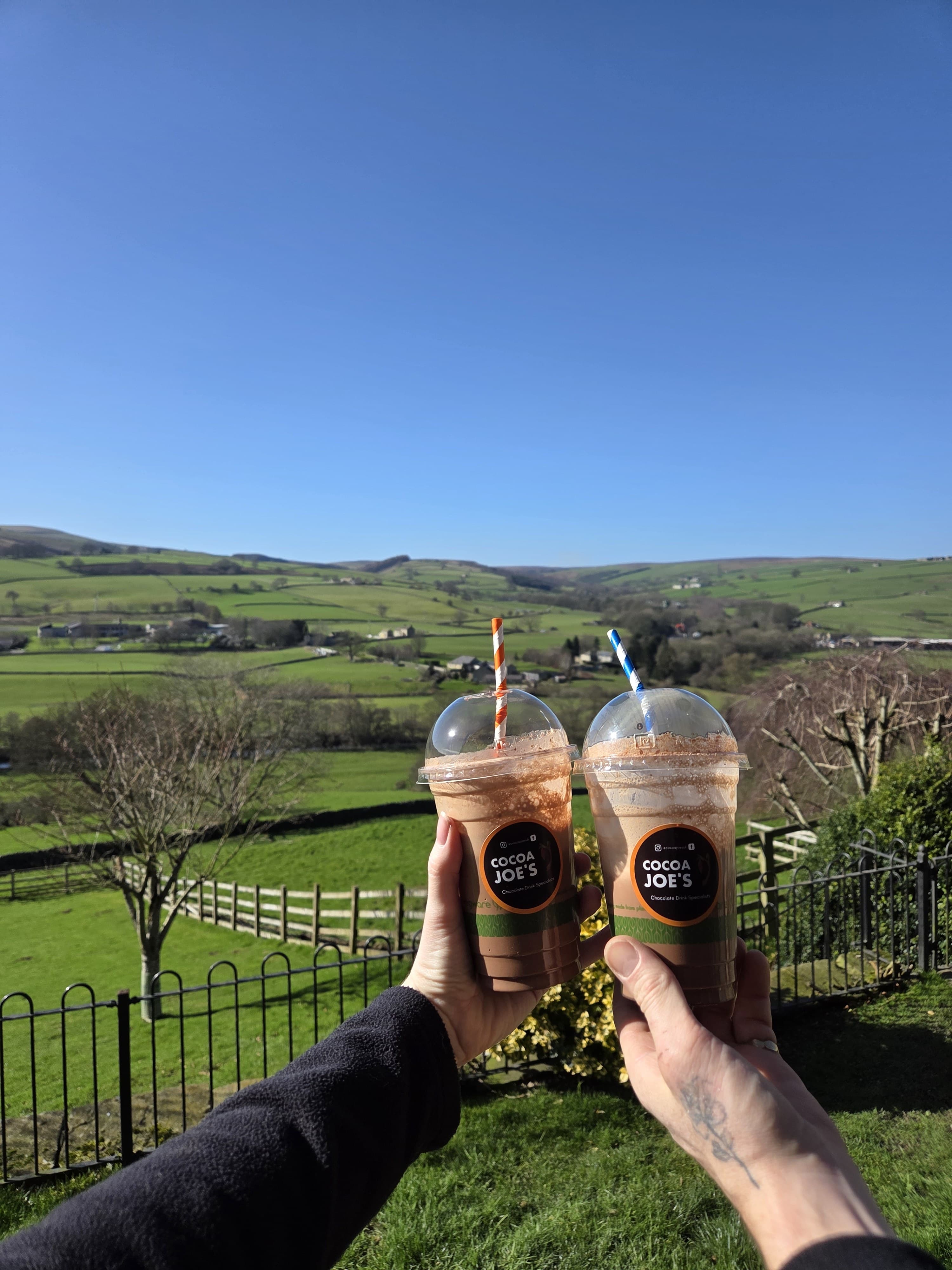 Two iced Cocoa Joe's drinks held against a wide blue-sky dale view in Nidderdale