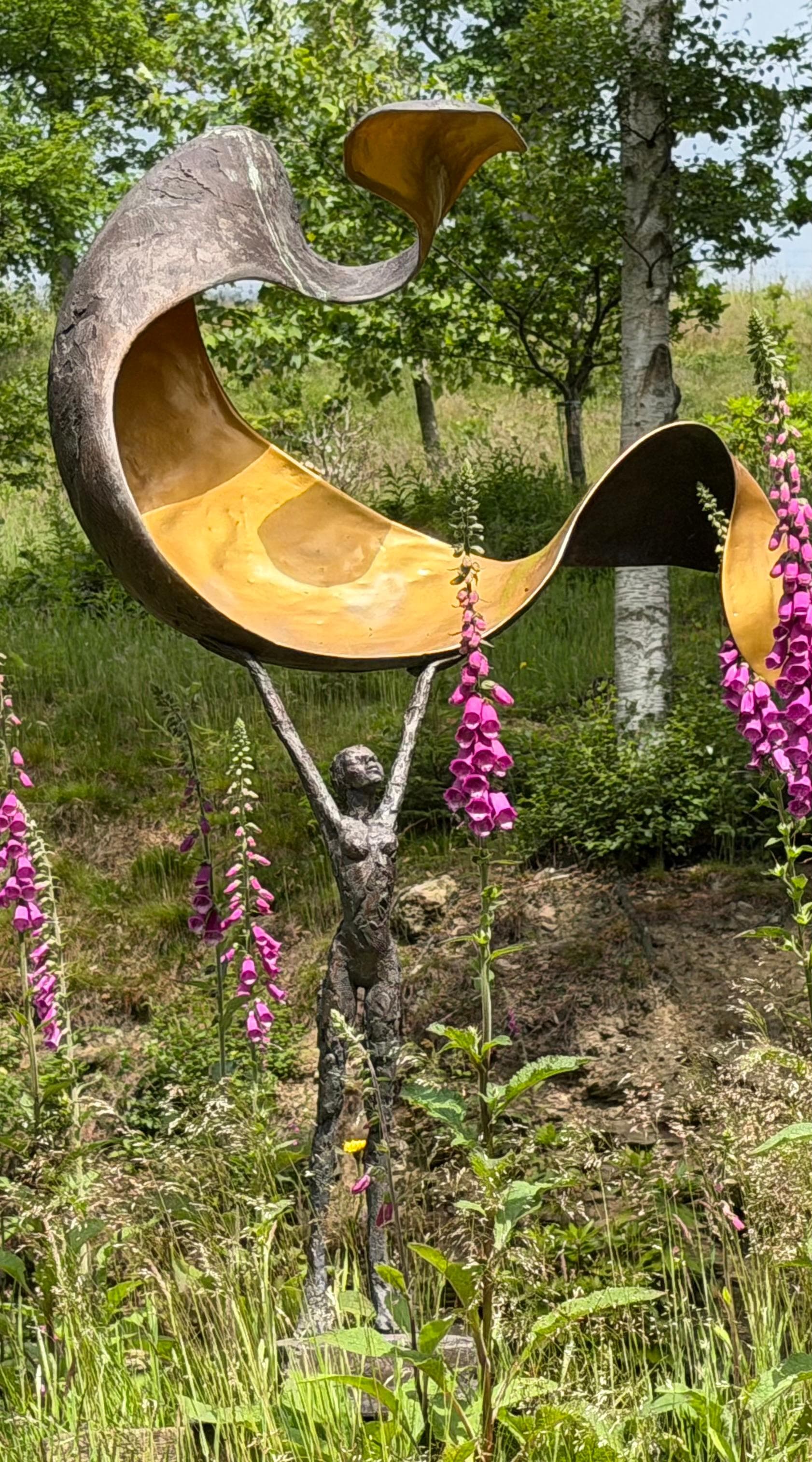 A bronze figure sculpture reaching up to a curved golden form, surrounded by pink foxgloves in the garden