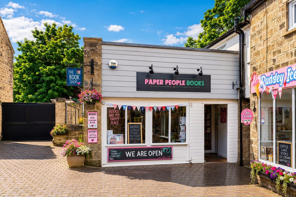 The white-fronted exterior of Paper People Books in Pateley Bridge, with bunting, hanging flower baskets and a chalkboard sign reading "We Are Open"