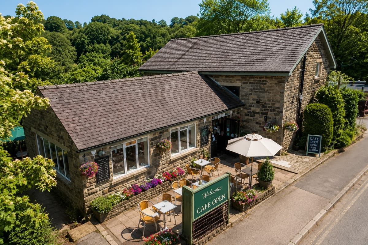 A stone cafe building with a terrace, parasol and flowers in bloom, surrounded by trees by the River Nidd
