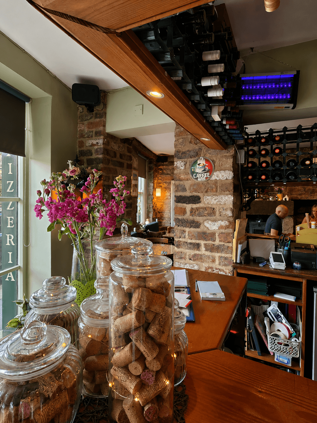 Fresh flowers and jars of wine corks on the bar at Olley's Pizzeria, with the kitchen pass and wine racks behind