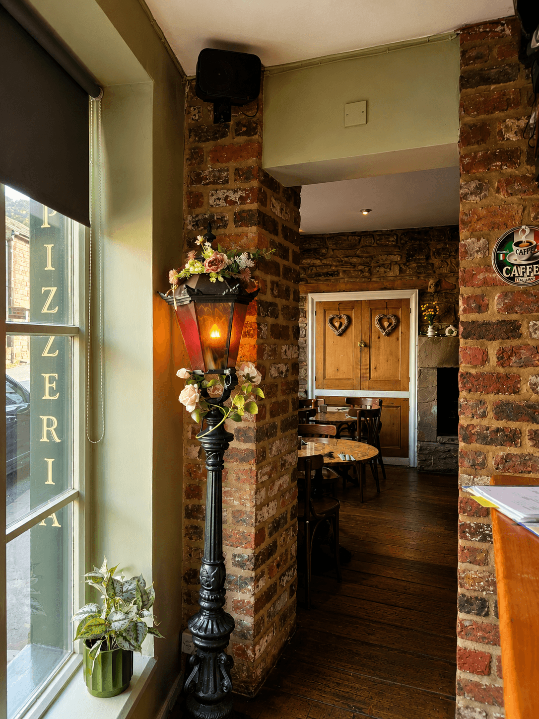 An ornate lamp post hung with flowers beside the brick archway into Olley's Pizzeria's dining room, with the PIZZERIA sign visible in the window